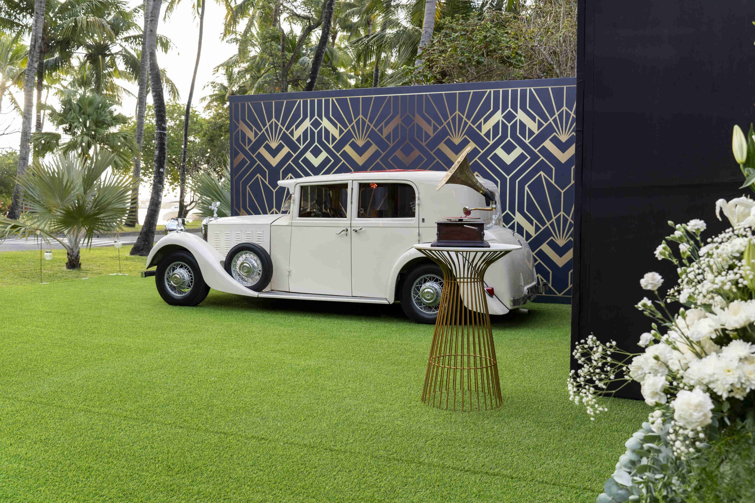 Vintage white car parked on lush green grass beside a geometric-patterned wall. A decorative table holds a record player, with flowers in the foreground.