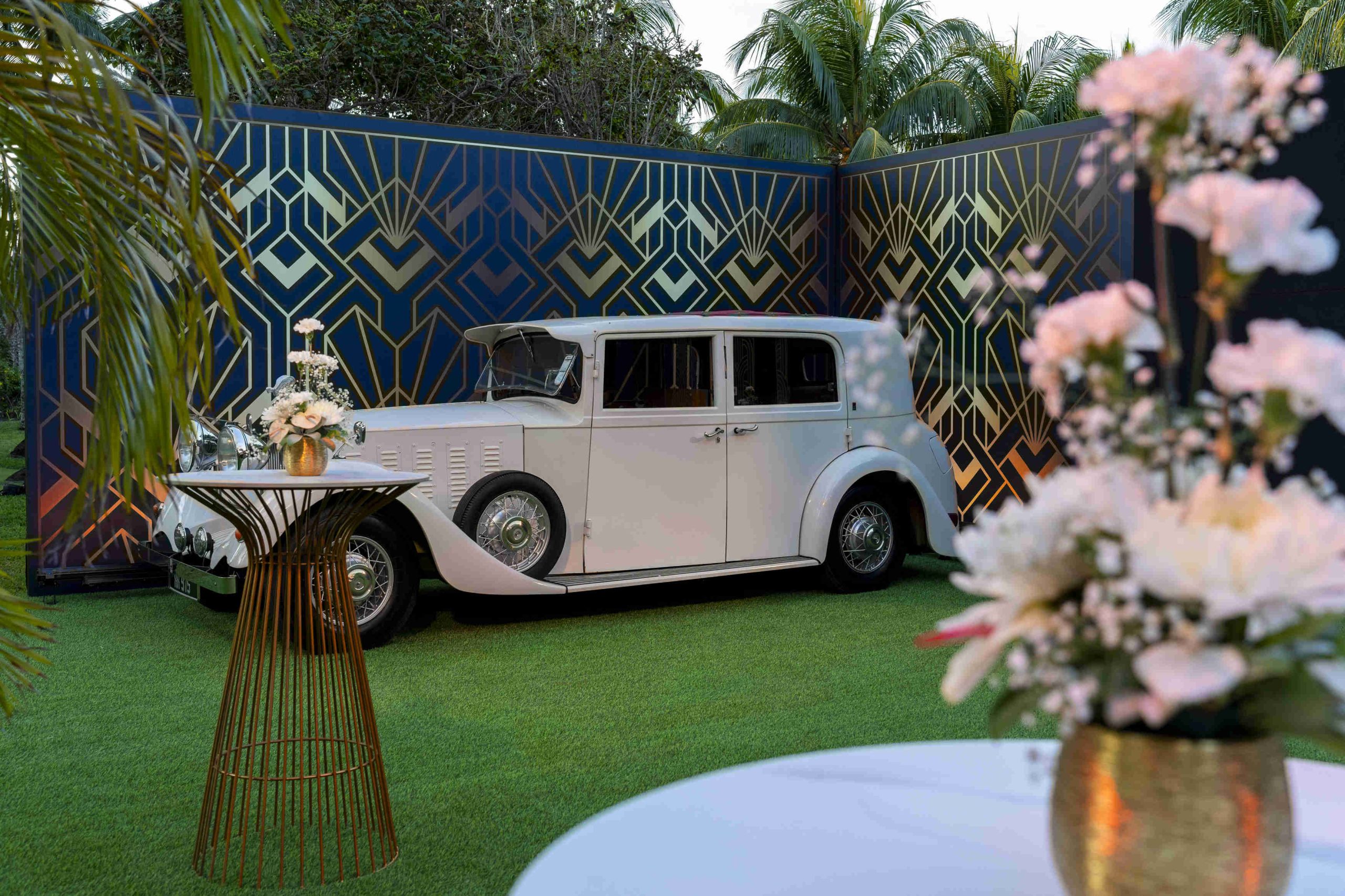 A vintage white car is parked on a lush green lawn, framed by elegant gold and navy art deco panels. Foreground floral arrangements add a touch of elegance.