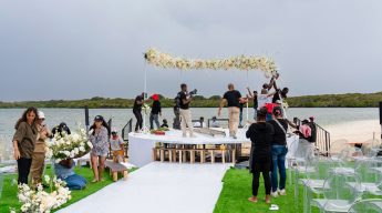 Event crew member smoothing the white floor covering on the circular waterfront stage set next to the water's edge, featuring a large floral arch, white lounge sofa, and chairs, with a natural landscape backdrop under a cloudy sky.