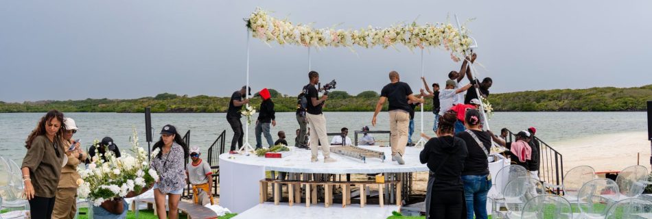 Event crew member smoothing the white floor covering on the circular waterfront stage set next to the water's edge, featuring a large floral arch, white lounge sofa, and chairs, with a natural landscape backdrop under a cloudy sky.