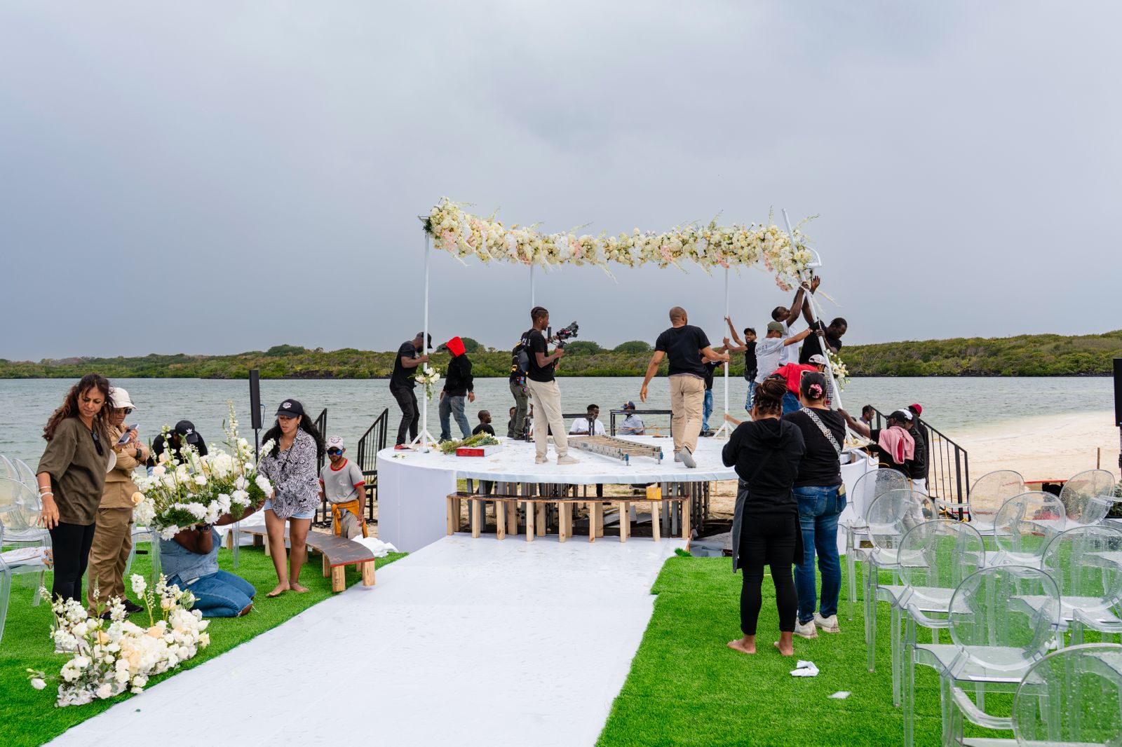 Event crew member smoothing the white floor covering on the circular waterfront stage set next to the water's edge, featuring a large floral arch, white lounge sofa, and chairs, with a natural landscape backdrop under a cloudy sky.