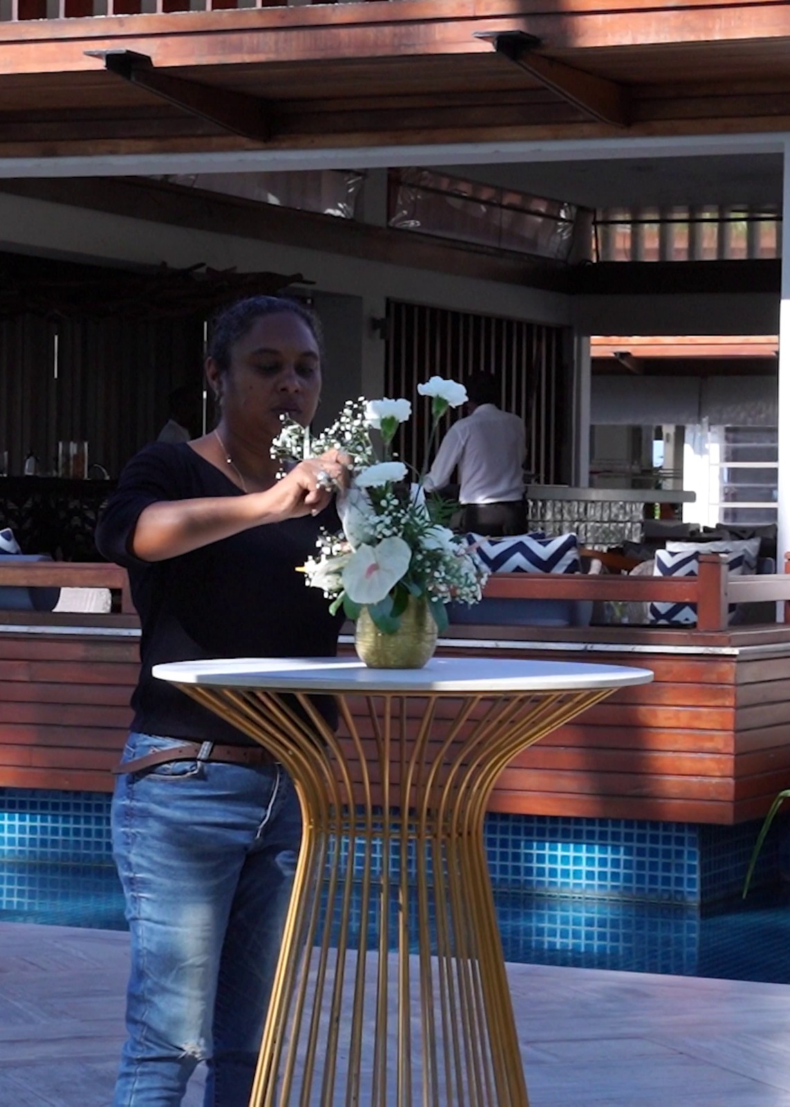 A woman in a black shirt and jeans arranges a floral centerpiece on a modern round table with a gold stand. The setting is a stylish, outdoor lounge area with blue accents.