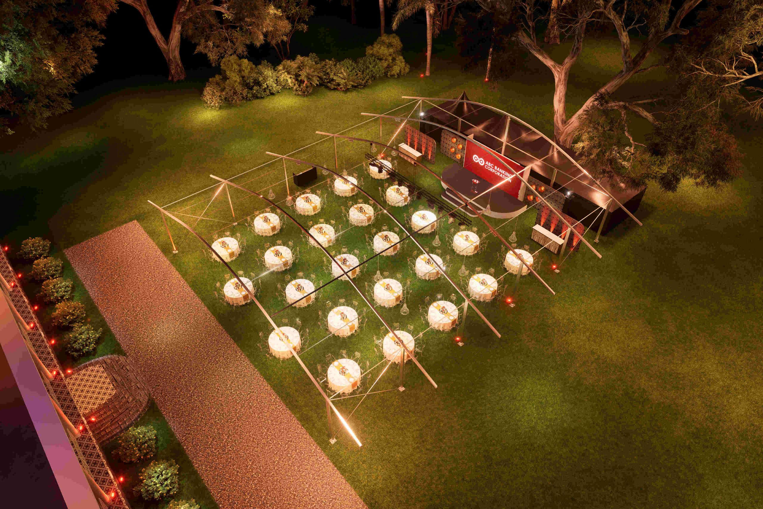 Outdoor event setup with circular tables lit under a canopy on a grassy area, string lights creating a warm ambiance; a stage to the right with trees behind.