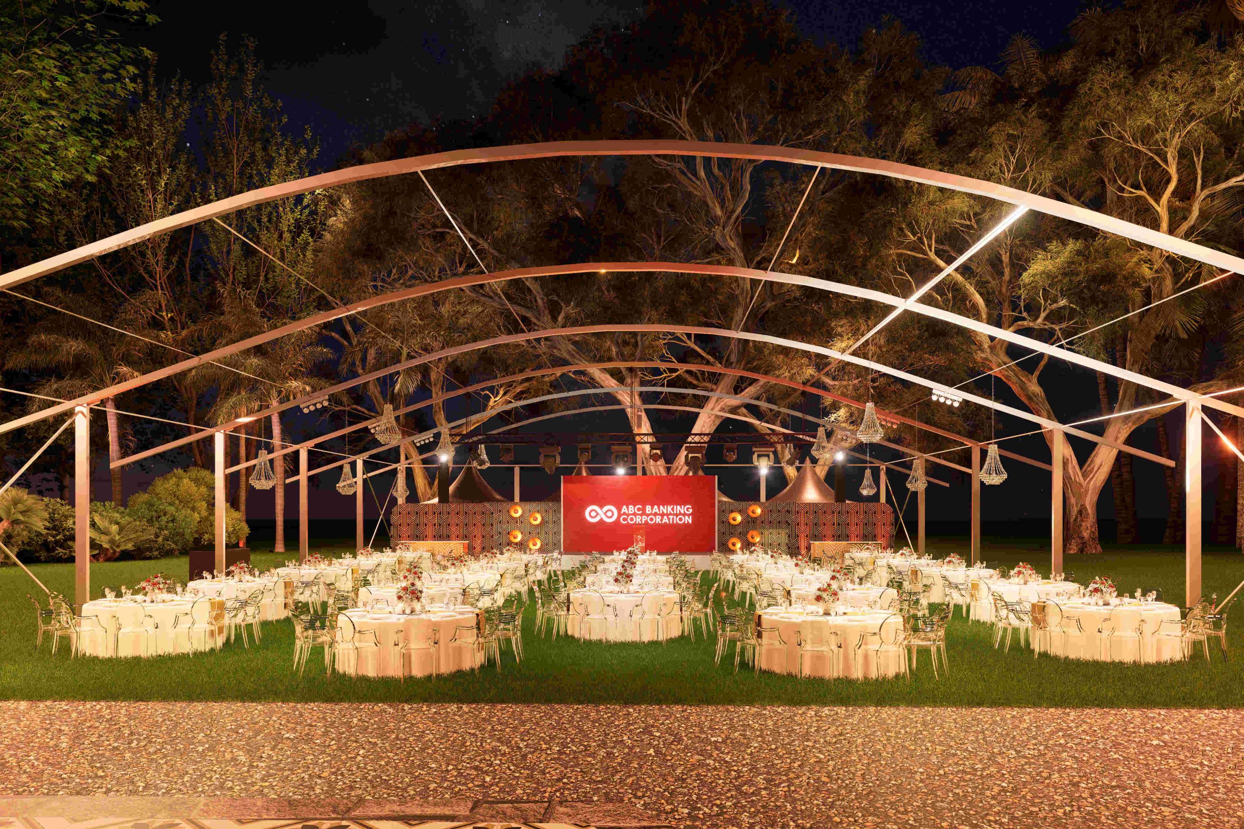 Outdoor event setup at night, featuring round tables with white tablecloths under a lit canopy. A corporate banner in the background reads “ABC Banking Corporation.” The scene is surrounded by trees, creating a formal and inviting atmosphere.