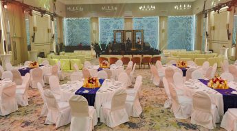 An elegant banquet hall set for an event, featuring round tables with white cloths and floral centerpieces. A stage with ornamental frames is in the background.