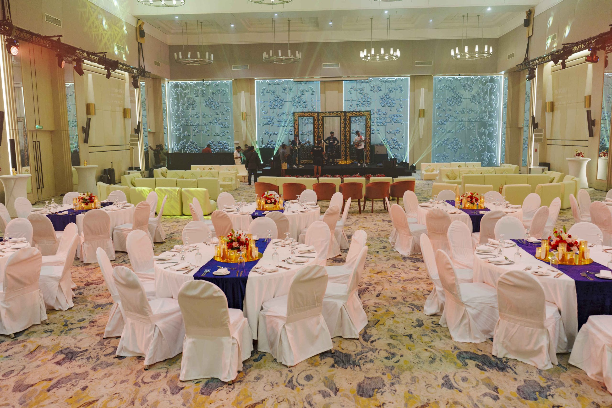 An elegant banquet hall set for an event, featuring round tables with white cloths and floral centerpieces. A stage with ornamental frames is in the background.