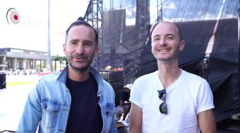 Two male members of the Glorious Artist group smiling in front of a large outdoor stage structure with the Impact Production Group logo visible.