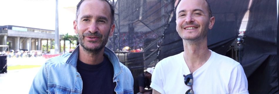 Two male members of the Glorious Artist group smiling in front of a large outdoor stage structure with the Impact Production Group logo visible.