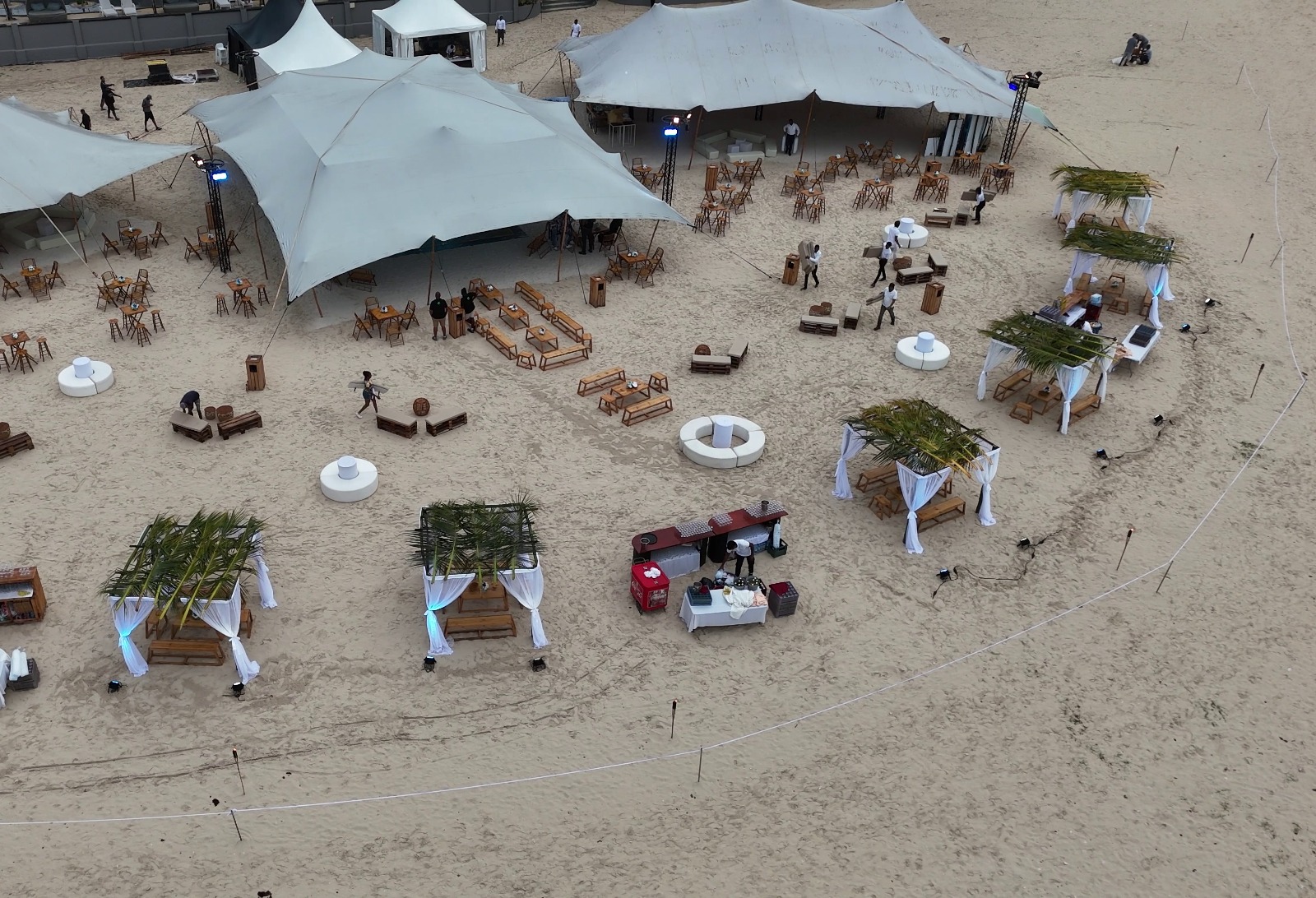 Aerial view of a beach event setup with large white tents, wooden furniture, and decorated canopies. People are arranging tables and chairs on the sand.