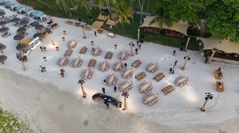 Aerial view of an elegant beach wedding setup. Rows of banquet tables on white sand are adorned with white tablecloths. Surrounded by palm trees, shaded lounge areas, and oceanfront.