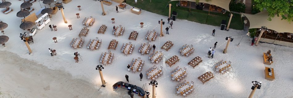 Aerial view of an elegant beach wedding setup. Rows of banquet tables on white sand are adorned with white tablecloths. Surrounded by palm trees, shaded lounge areas, and oceanfront.