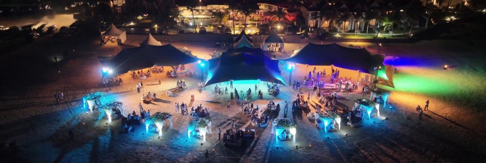 Aerial view of a vibrant nighttime beach party with colorful lights illuminating large tents and scattered groups of people, set against a lively town backdrop.