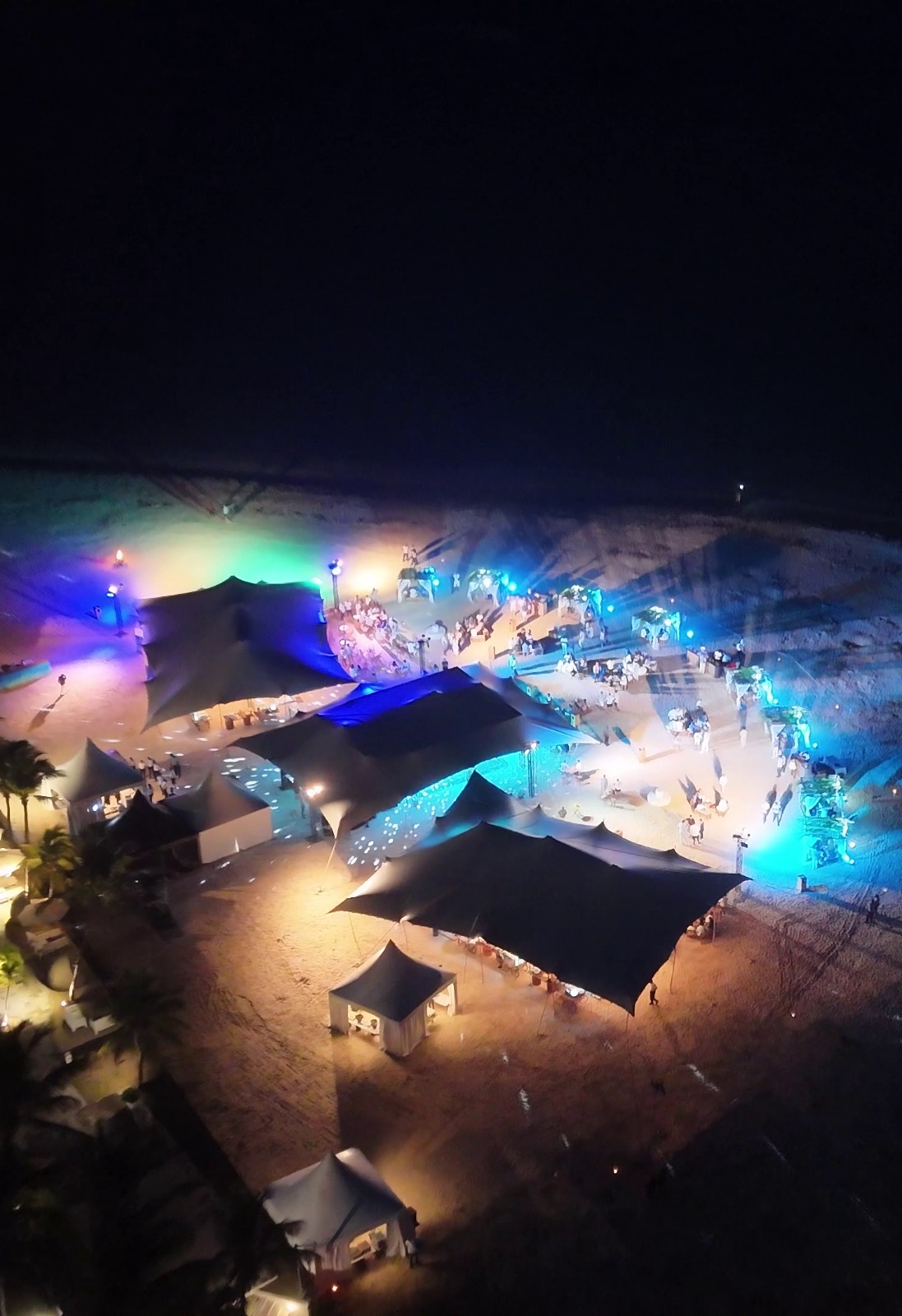 Aerial view of a vibrant beach party at night, featuring colorful tents illuminated in blue and purple lights with scattered groups of people.