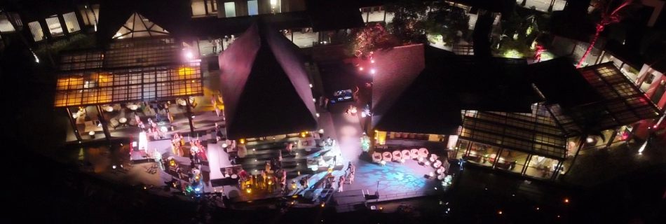Aerial view of a night event at a resort with illuminated pathways. Guests gather around brightly lit tables, evoking a lively and festive atmosphere.