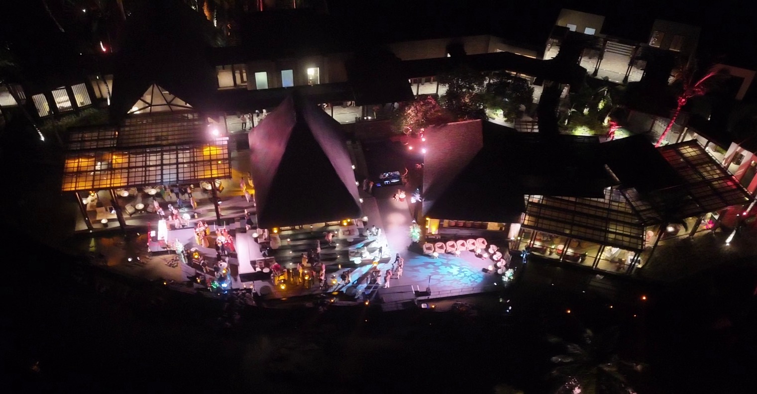 Aerial view of a night event at a resort with illuminated pathways. Guests gather around brightly lit tables, evoking a lively and festive atmosphere.