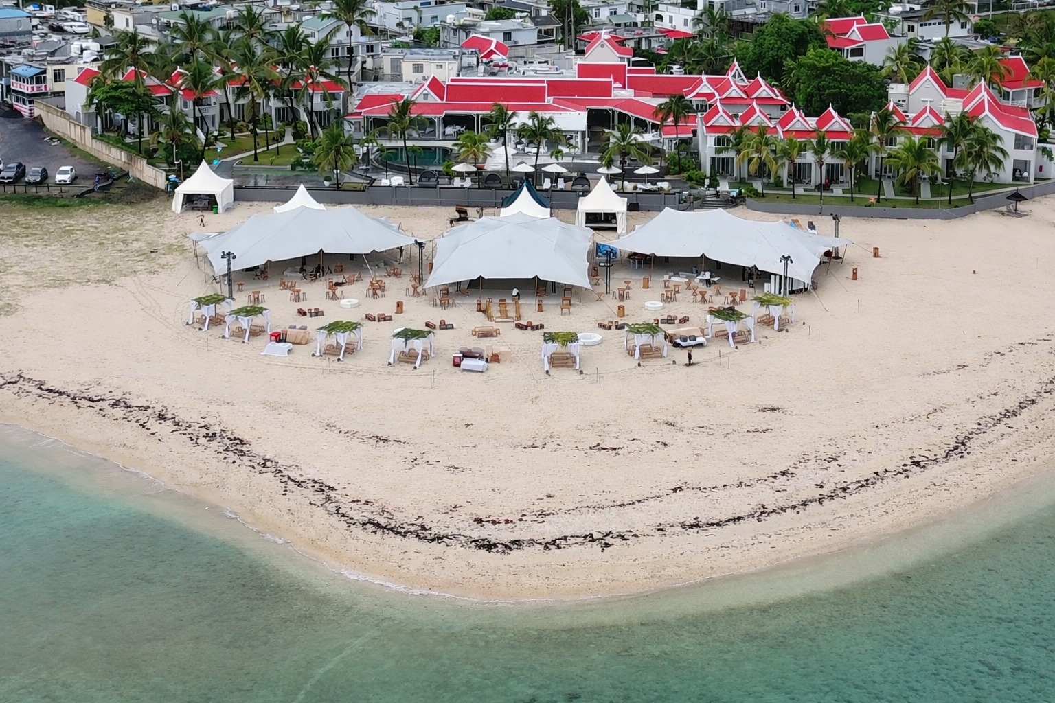 Aerial view of a pristine beach with white tents and lounge areas, set against a backdrop of vibrant red-roofed buildings and lush palm trees, conveying a tranquil, tropical resort atmosphere.