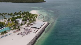 Aerial view of a tropical beach with palm trees and a large, white canopy covering a gathering of people on the sand. Turquoise ocean surrounds the area.