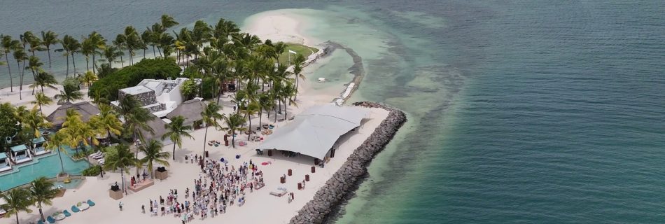 Aerial view of a tropical beach with palm trees and a large, white canopy covering a gathering of people on the sand. Turquoise ocean surrounds the area.