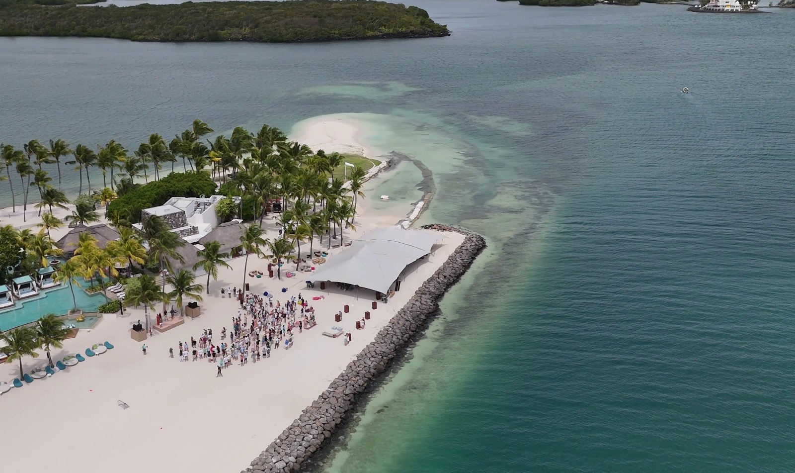 Aerial view of a tropical beach with palm trees and a large, white canopy covering a gathering of people on the sand. Turquoise ocean surrounds the area.