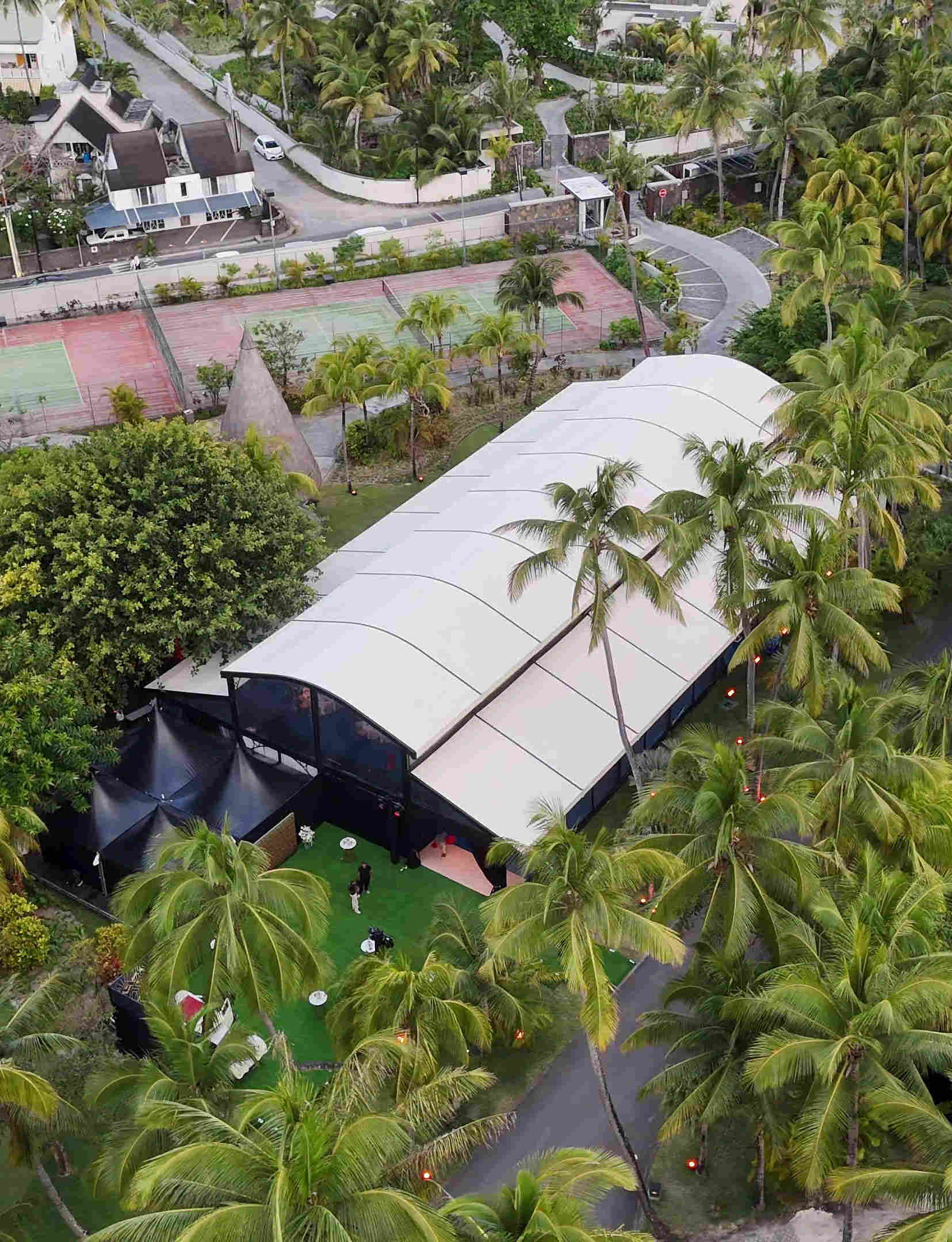 Aerial view of a tropical outdoor venue surrounded by palm trees, featuring a large tent with a white roof, nearby tennis courts, and a winding road. The scene conveys a serene, lush setting.