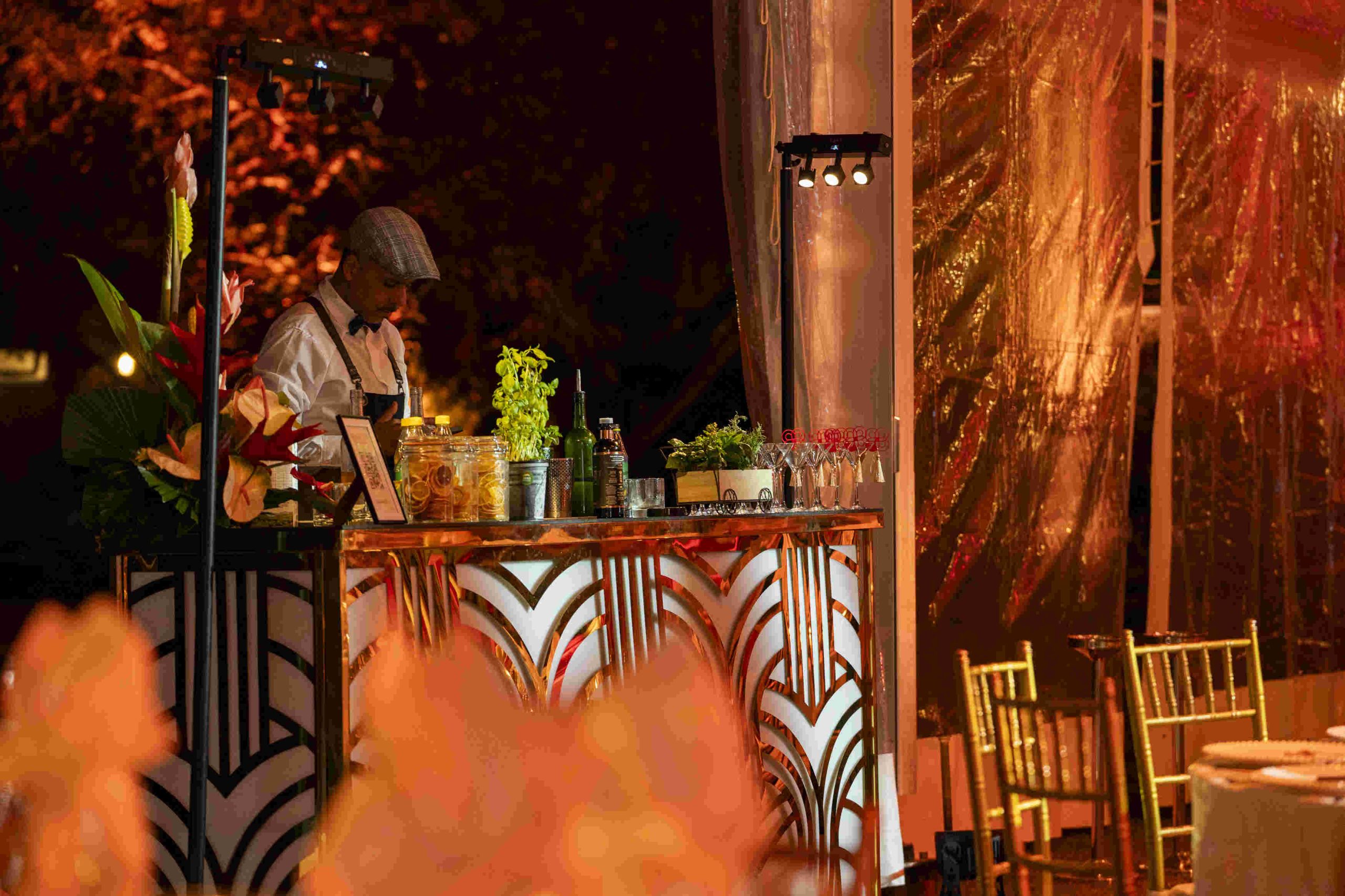 A bartender at a decorated outdoor bar mixes drinks under warm lights. The setup features floral arrangements, greenery, and elegant golden chairs nearby.