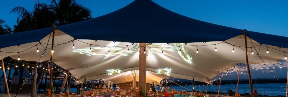 A beachside event setup at dusk with a large canopy tent lit by string lights. Tables are adorned with bright tablecloths and floral centerpieces, creating a festive, tropical ambiance.