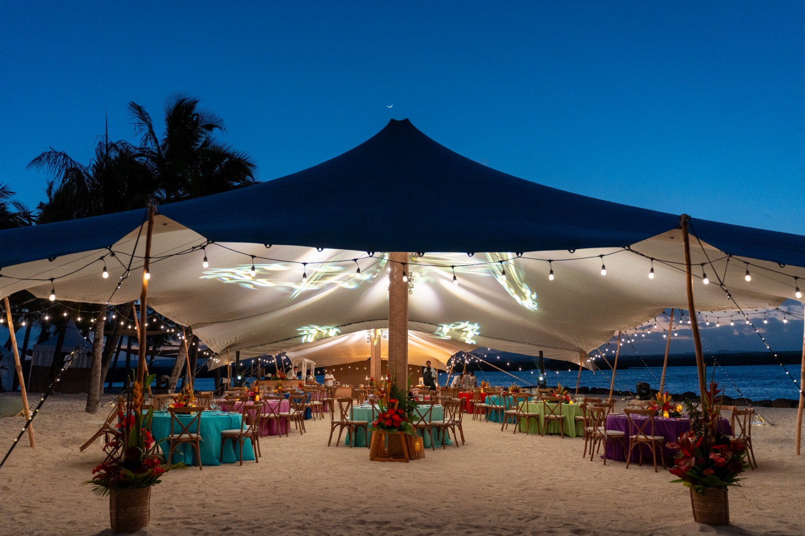 A beachside event setup at dusk with a large canopy tent lit by string lights. Tables are adorned with bright tablecloths and floral centerpieces, creating a festive, tropical ambiance.