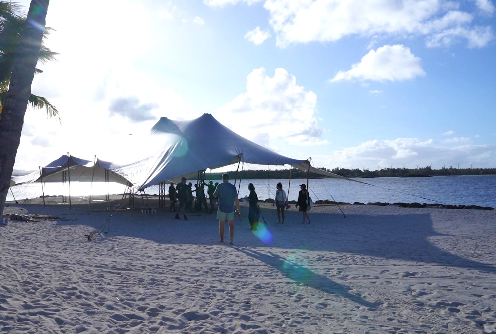 Outdoor beach event at sunset with a white canopy tent and silhouetted people beneath. The sky is bright blue with scattered clouds, creating a serene ambiance.