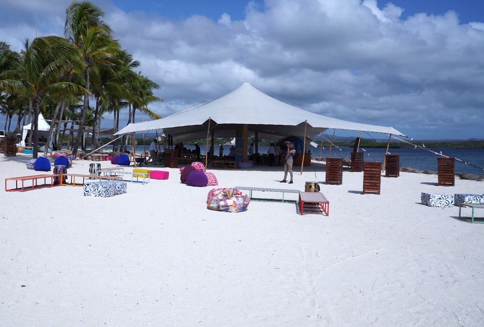 Outdoor beach event with a large white tent, colorful bean bags, and wooden tables on sand. Palm trees and ocean in the background under cloudy skies.