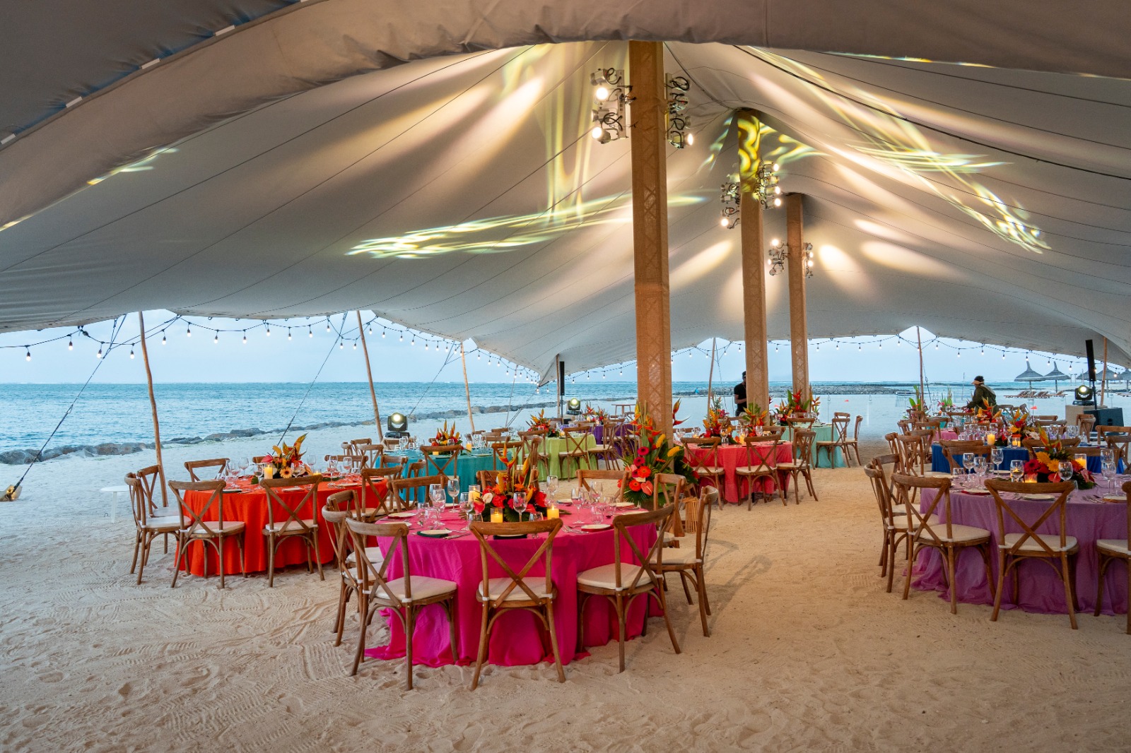 A festive beach event under a large white tent with round tables draped in colorful cloths. Warm lighting creates a welcoming ambiance near the ocean.