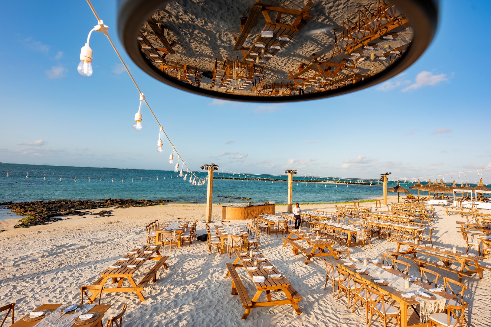 Beachside event setup with wooden tables and chairs on sand near clear blue water. String lights hang above, reflecting in a circular mirror for a festive feel.