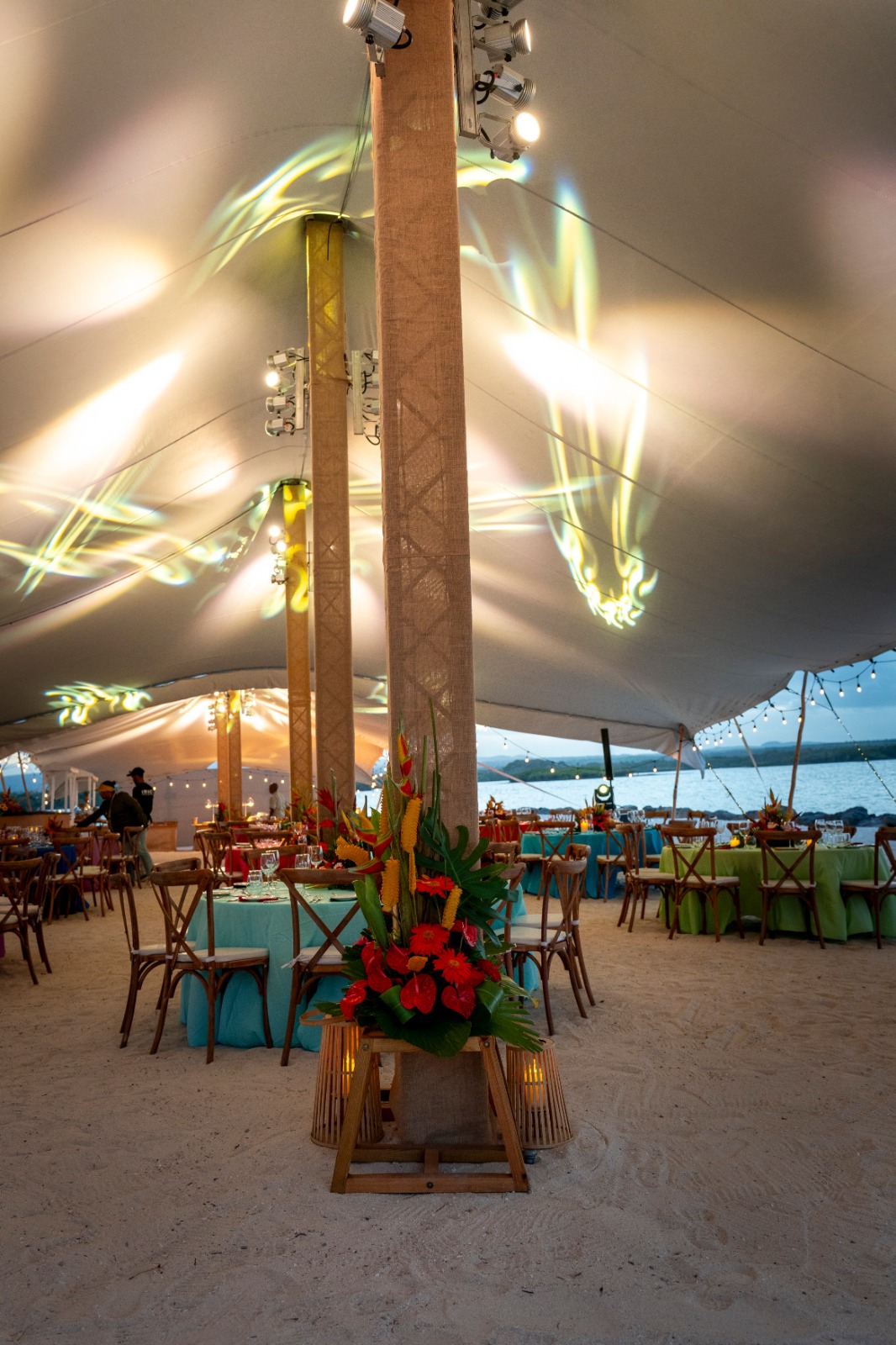 A beachside event tent adorned with colorful lighting and floral arrangements. Tables are set with turquoise and green linens, creating a festive ambiance.