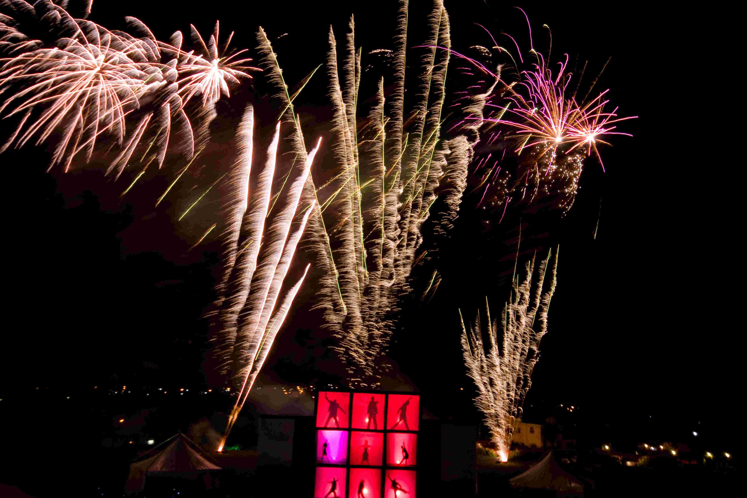 Bright fireworks explode in a night sky, casting colorful lights over silhouetted figures in red-lit windows. Festive and dynamic atmosphere.