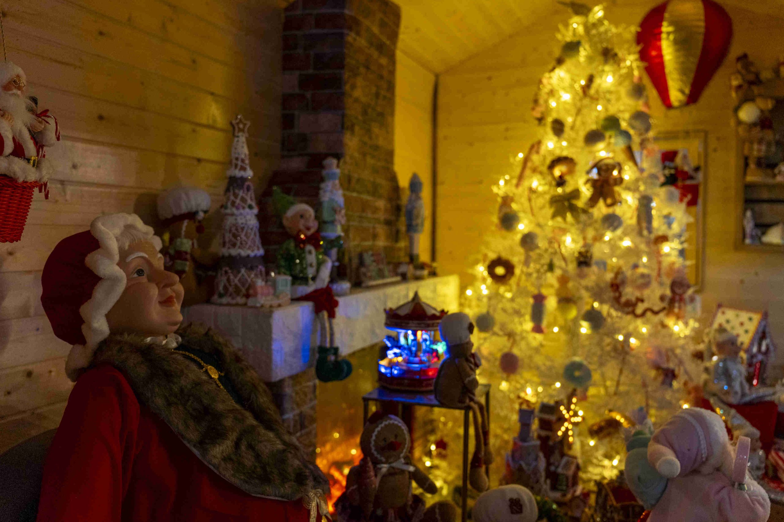Cozy Christmas room with warm lighting. A decorated tree glows with ornaments. A festive figure in red sits near a fireplace adorned with holiday decor.