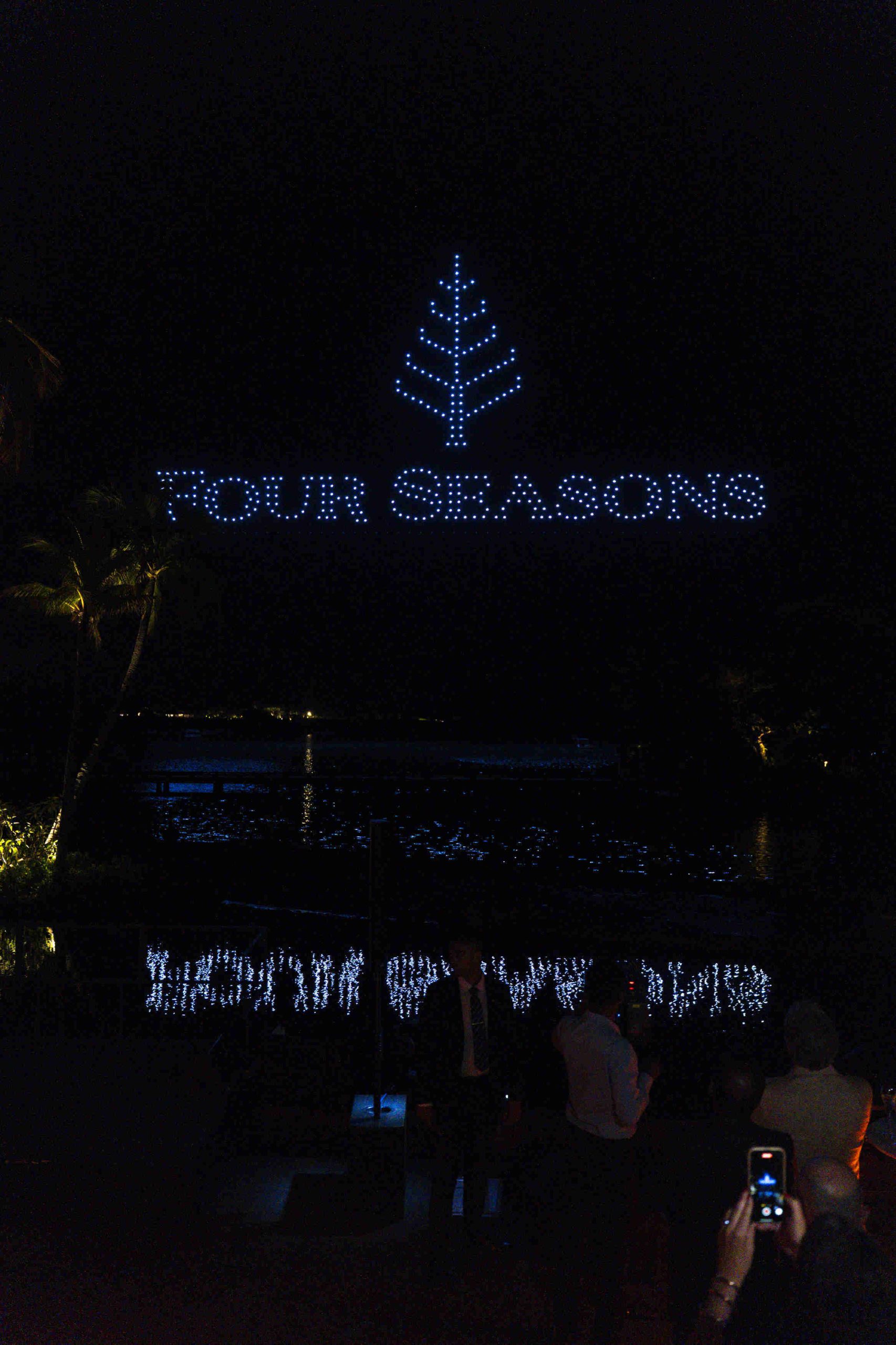 A drone light show displays the "Four Seasons" logo with a tree symbol above water at night, creating a reflective, enchanting, and festive atmosphere.