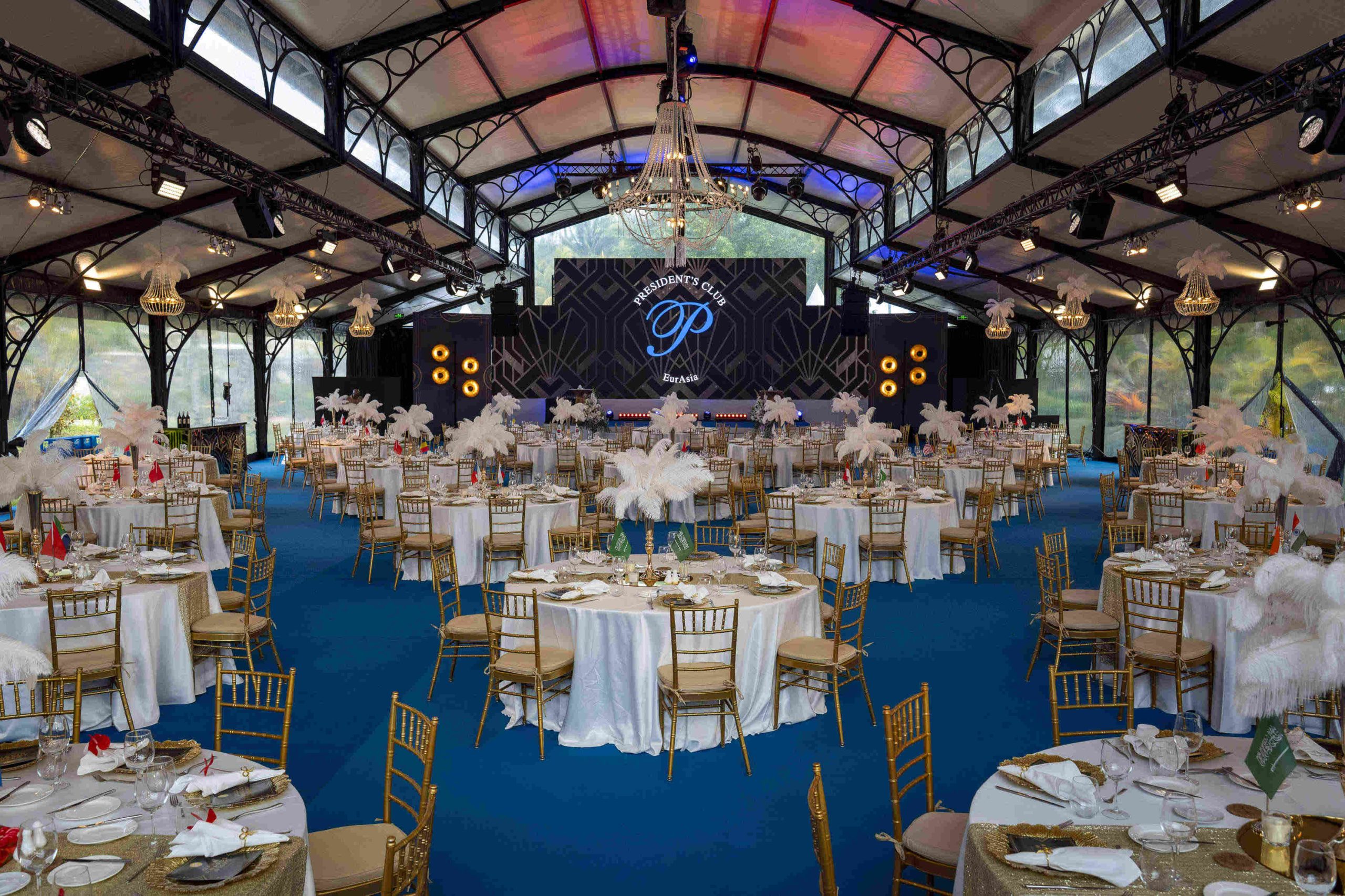 Elegant banquet hall with round tables, white linens, gold chairs, feather centerpieces, and a large stage. A chandelier hangs above, creating a lavish atmosphere.