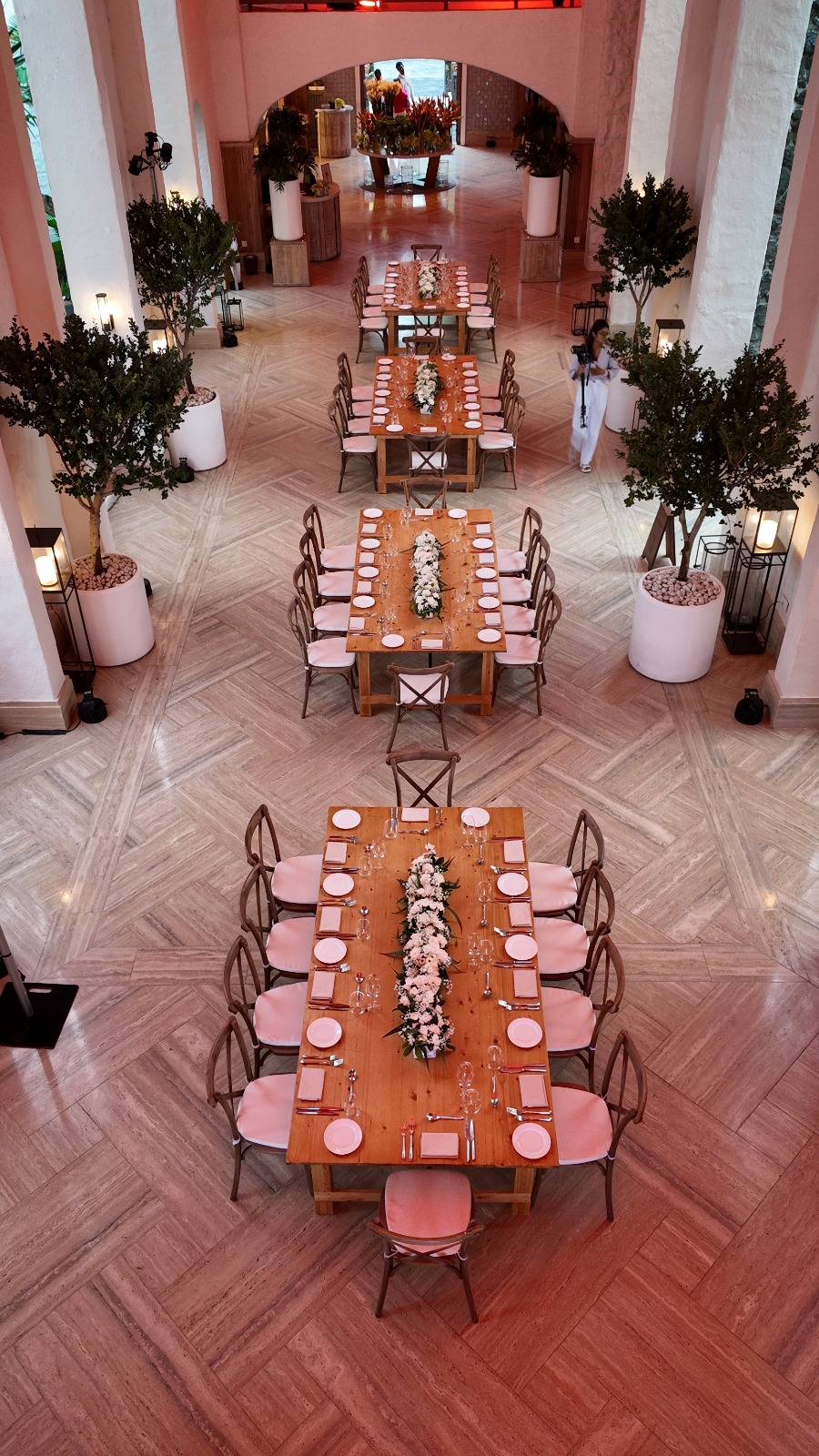 Elegant dining setup in a spacious hall with three wooden tables lined with white floral centerpieces. Warm lighting and potted trees create a cozy ambiance.