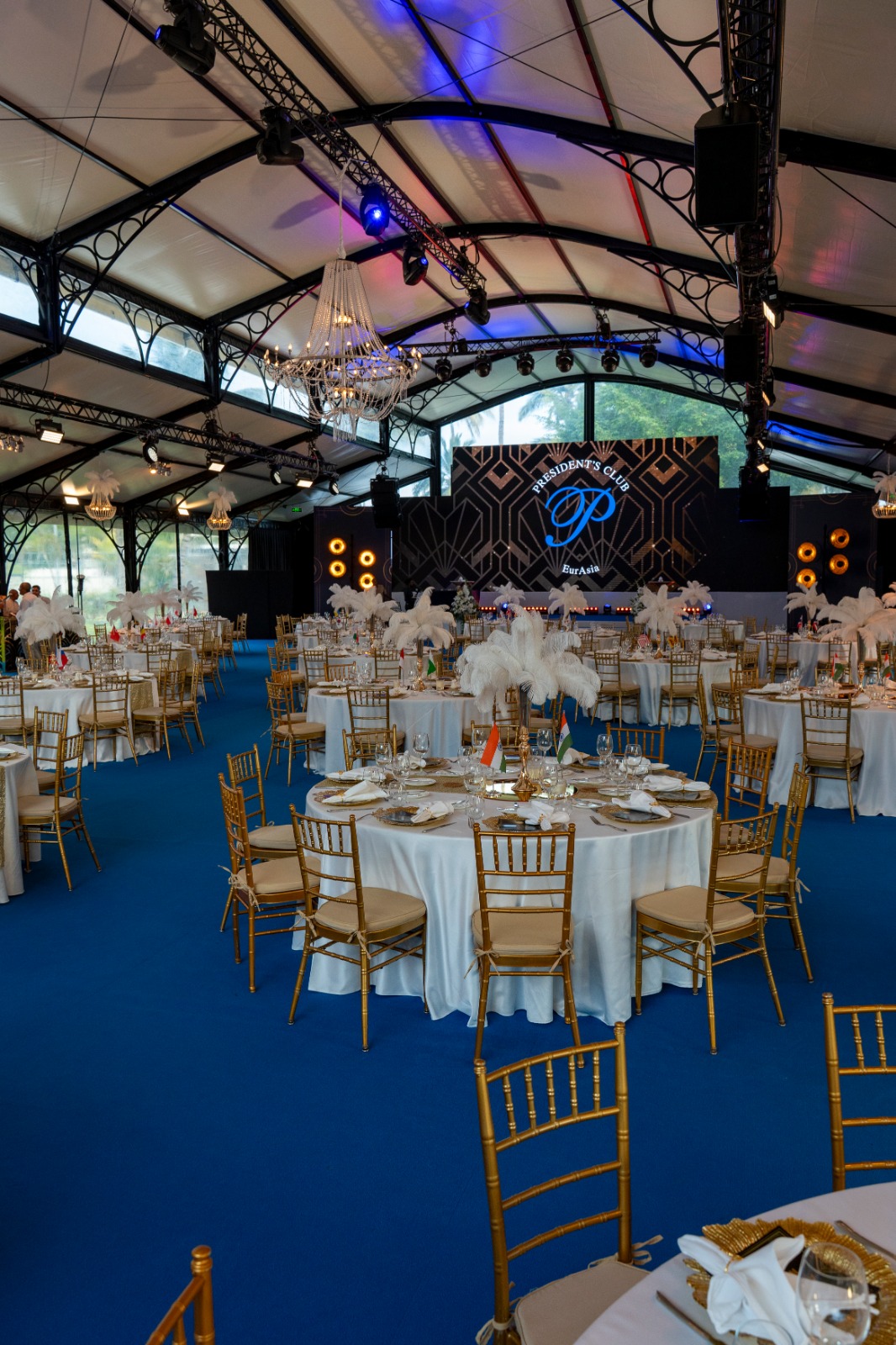 Elegant event hall with white-draped tables, gold chairs, and feather centerpieces on blue carpet. Chandelier lighting and geometric stage display. Festive ambiance.