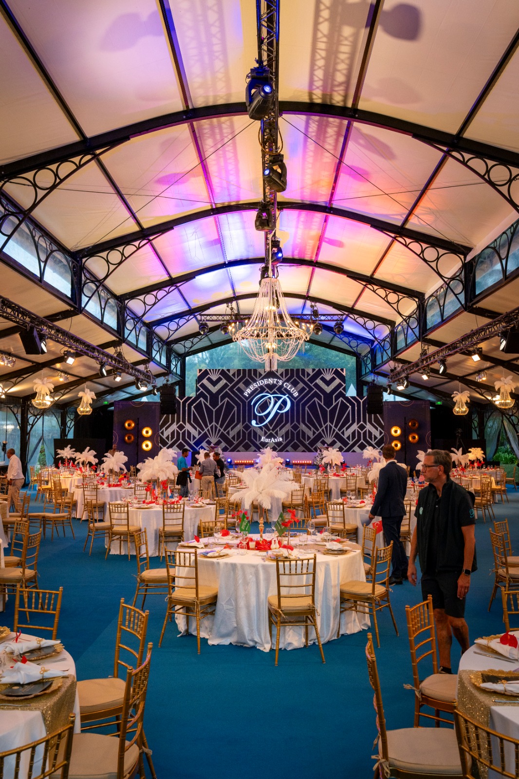 Elegant event hall with a chandelier, blue-lit ceiling, and art deco backdrop. Tables feature white and gold decor, promoting a festive ambiance.