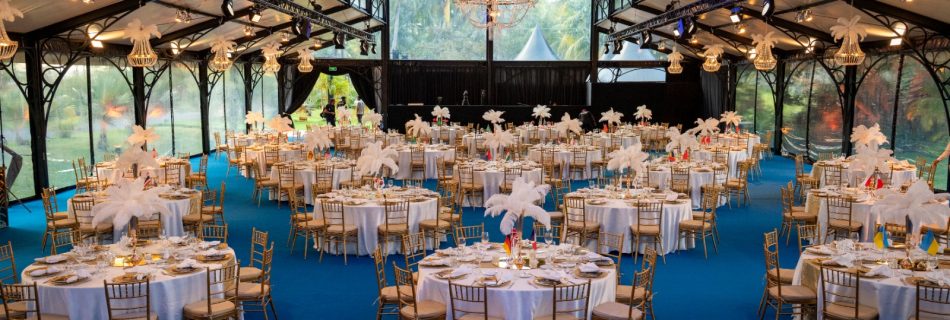 Elegant event space with blue carpet, round tables adorned with white tablecloths and feather centerpieces. Gold chairs add a touch of luxury.