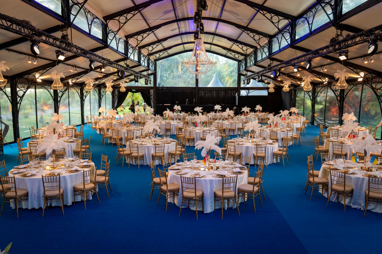 Elegant event space with blue carpet, round tables adorned with white tablecloths and feather centerpieces. Gold chairs add a touch of luxury.
