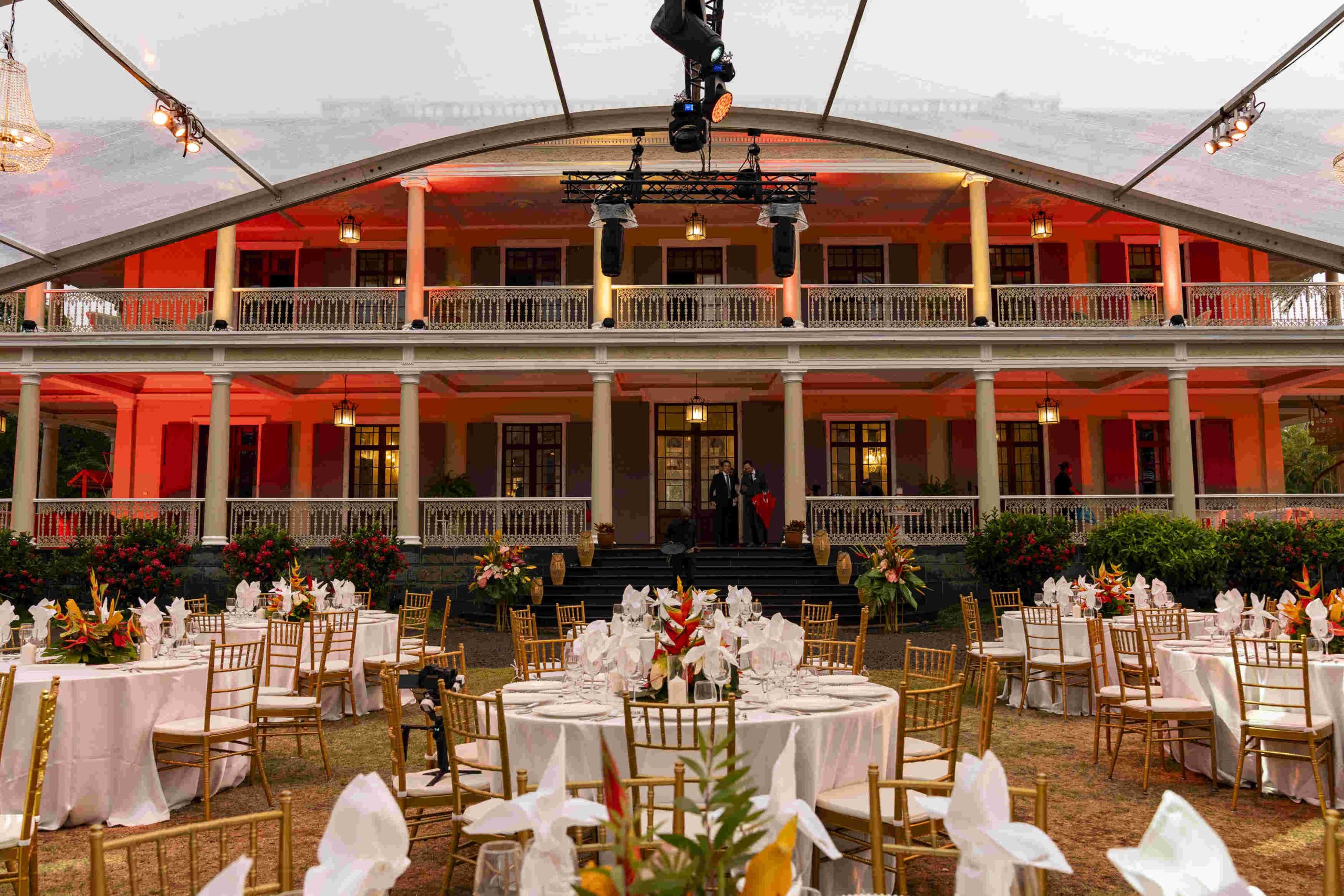 Elegant outdoor event with round tables, white linens, gold chairs, and floral centerpieces in front of a large, two-story building under tent lighting.