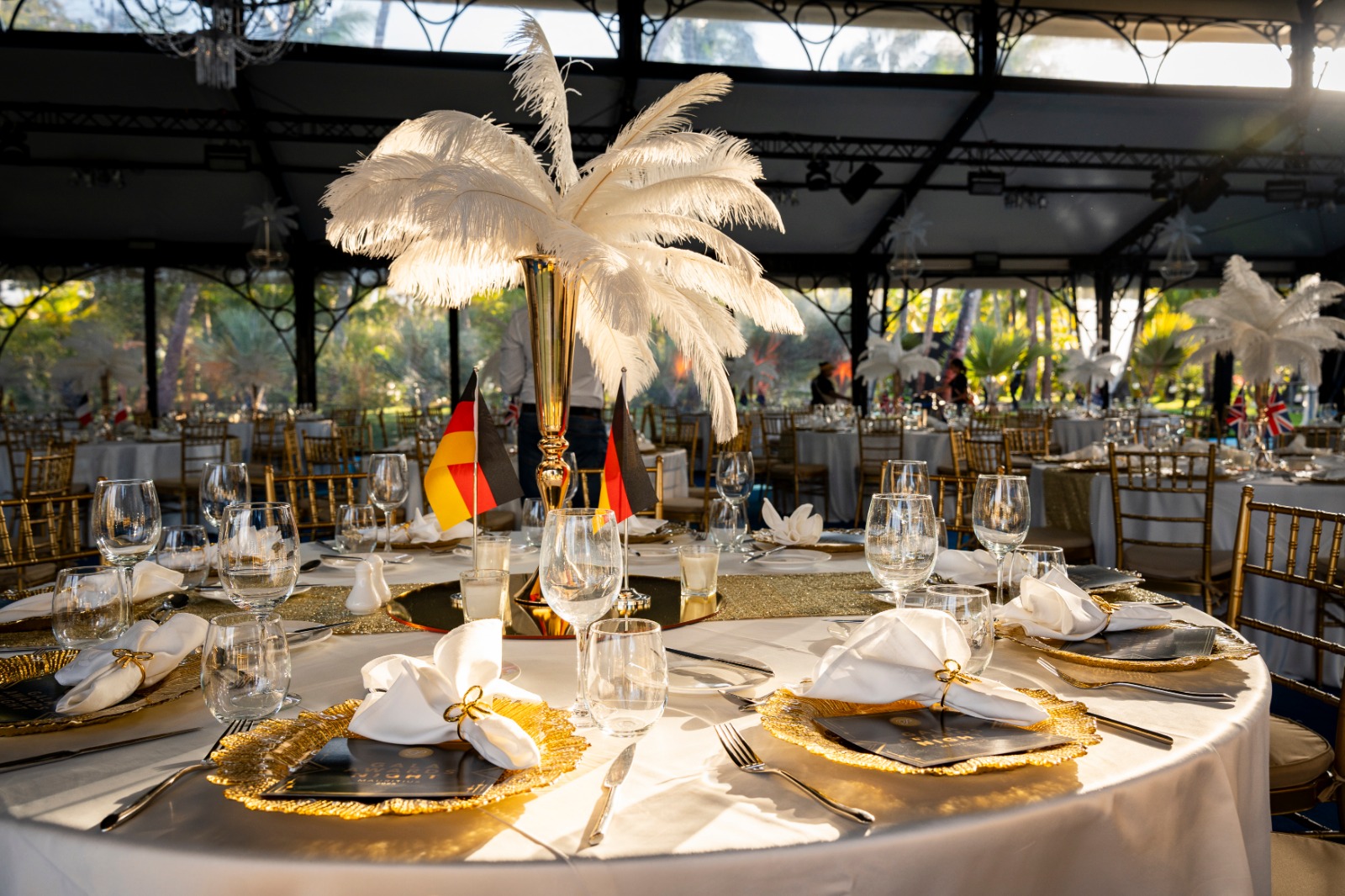 Elegant table setting with tall vase of white feathers at the center, surrounded by gold-trimmed plates, folded napkins, flags, and wine glasses. Luxurious ambiance.