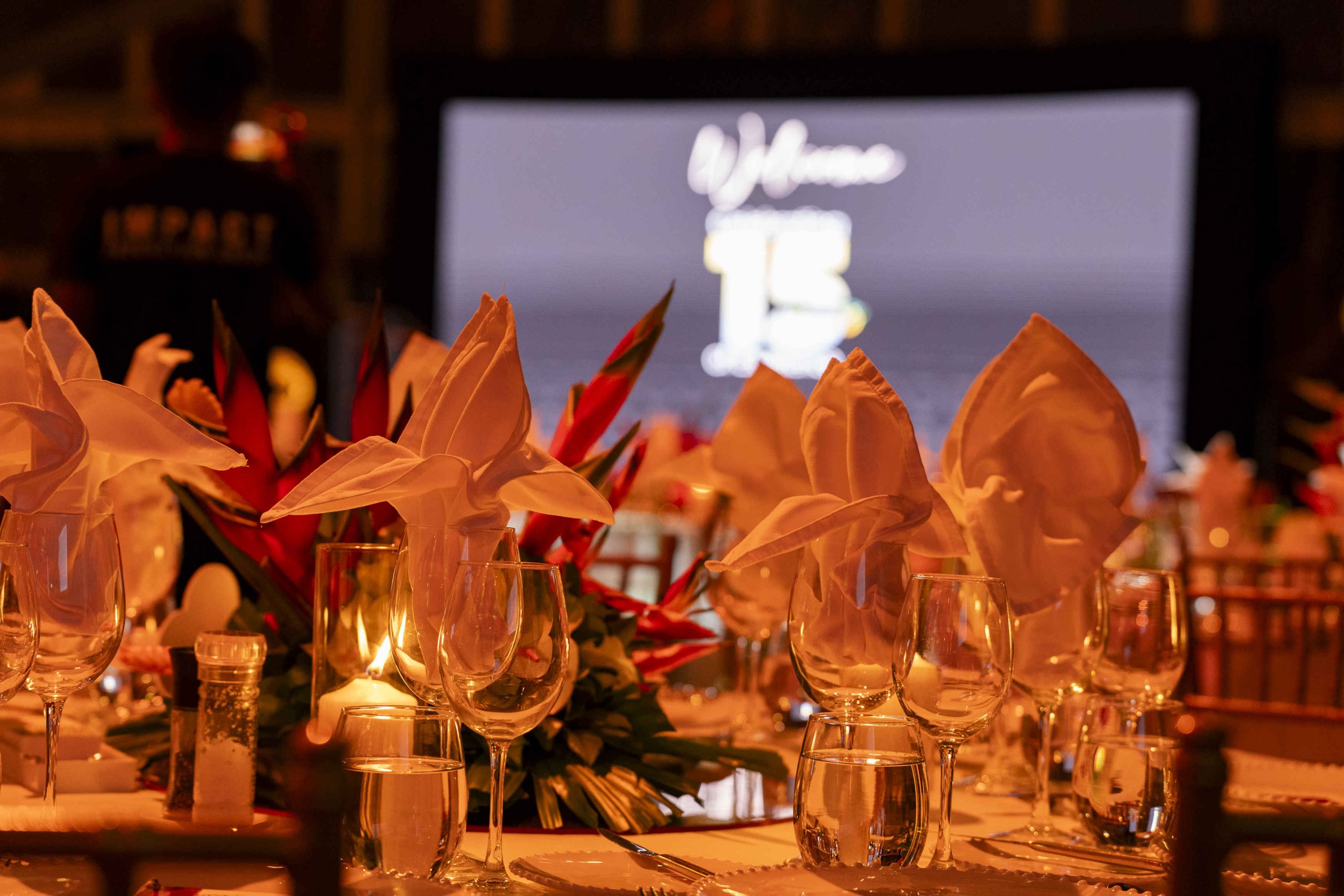 Elegant table setting with neatly folded white napkins, lit candles, and tropical flowers. A blurry screen in the background exudes a warm, festive ambiance.