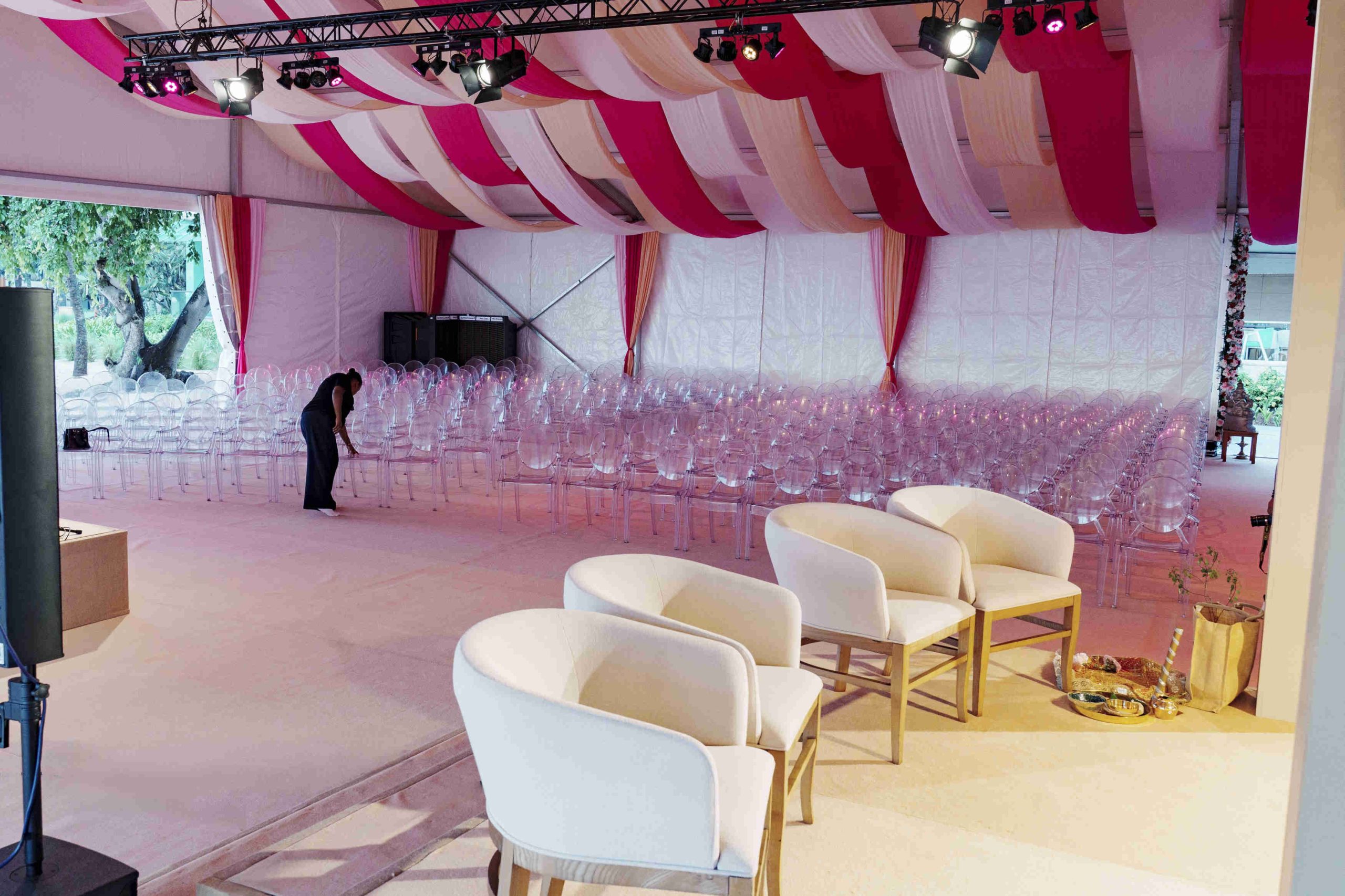 Spacious, elegantly decorated venue with red and white drapes, transparent chairs, and white armchairs. A person adjusts seating, creating a formal ambiance.