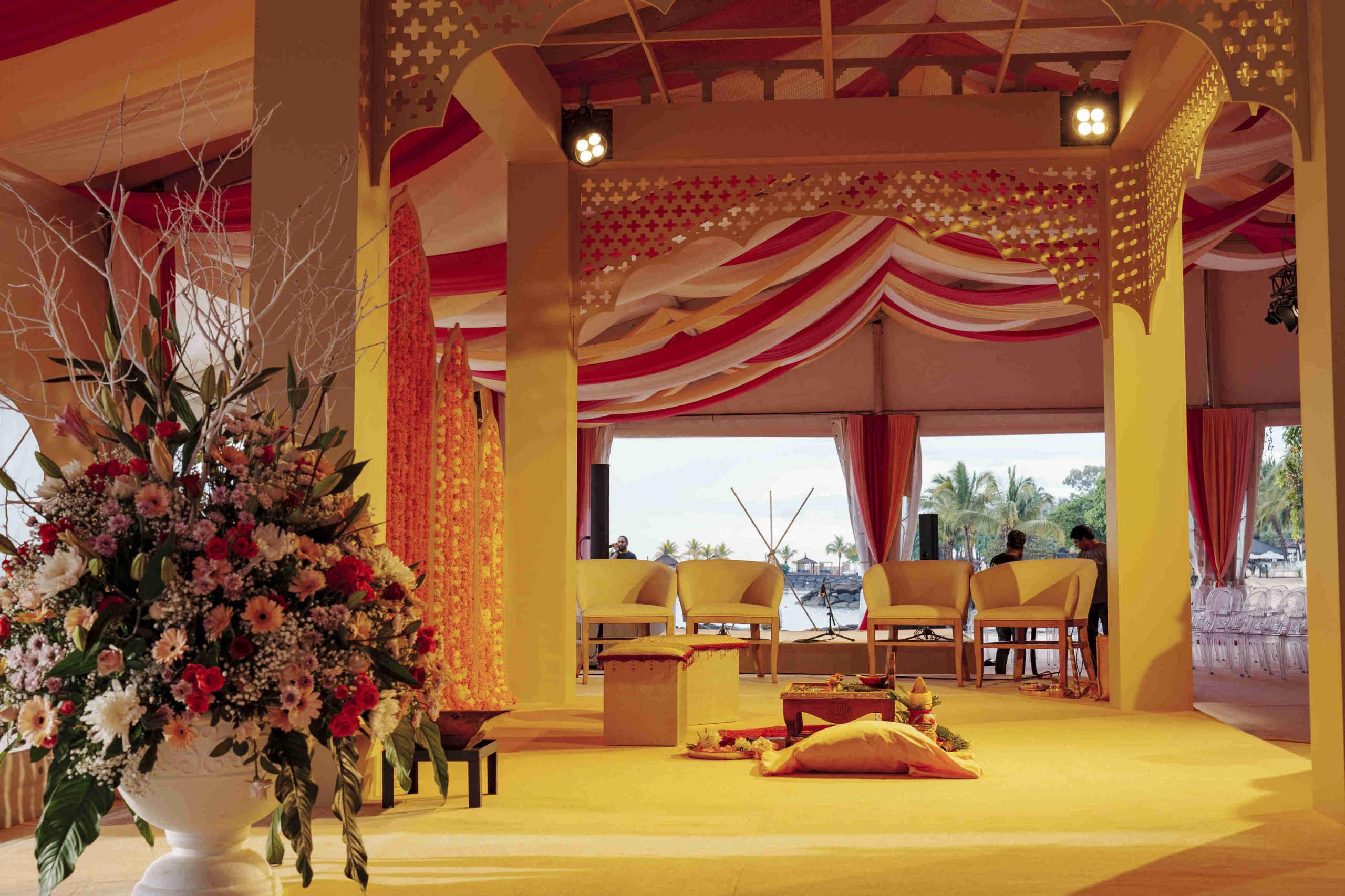 Spacious event space with elegant red and white drapery, a large floral arrangement on the left, and cream chairs arranged facing waterfront views.