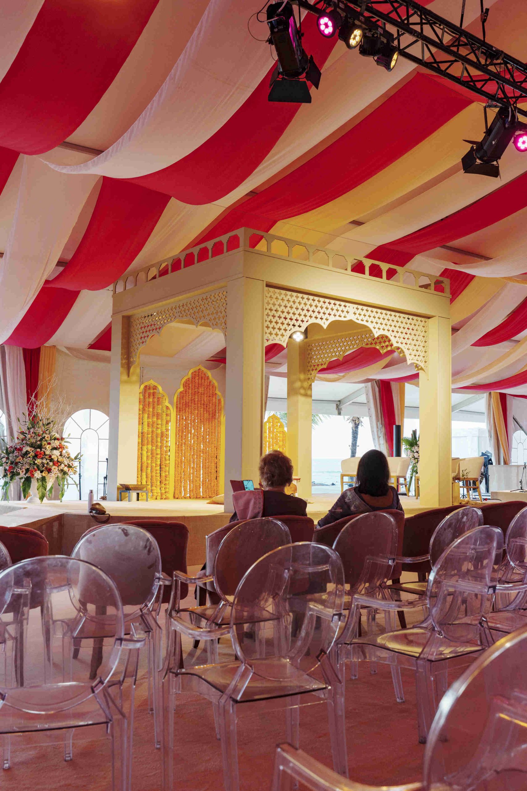 A vibrant, decorated event space with red and white drapery, golden ornate stage, transparent chairs, and floral arrangements, conveying a festive atmosphere.