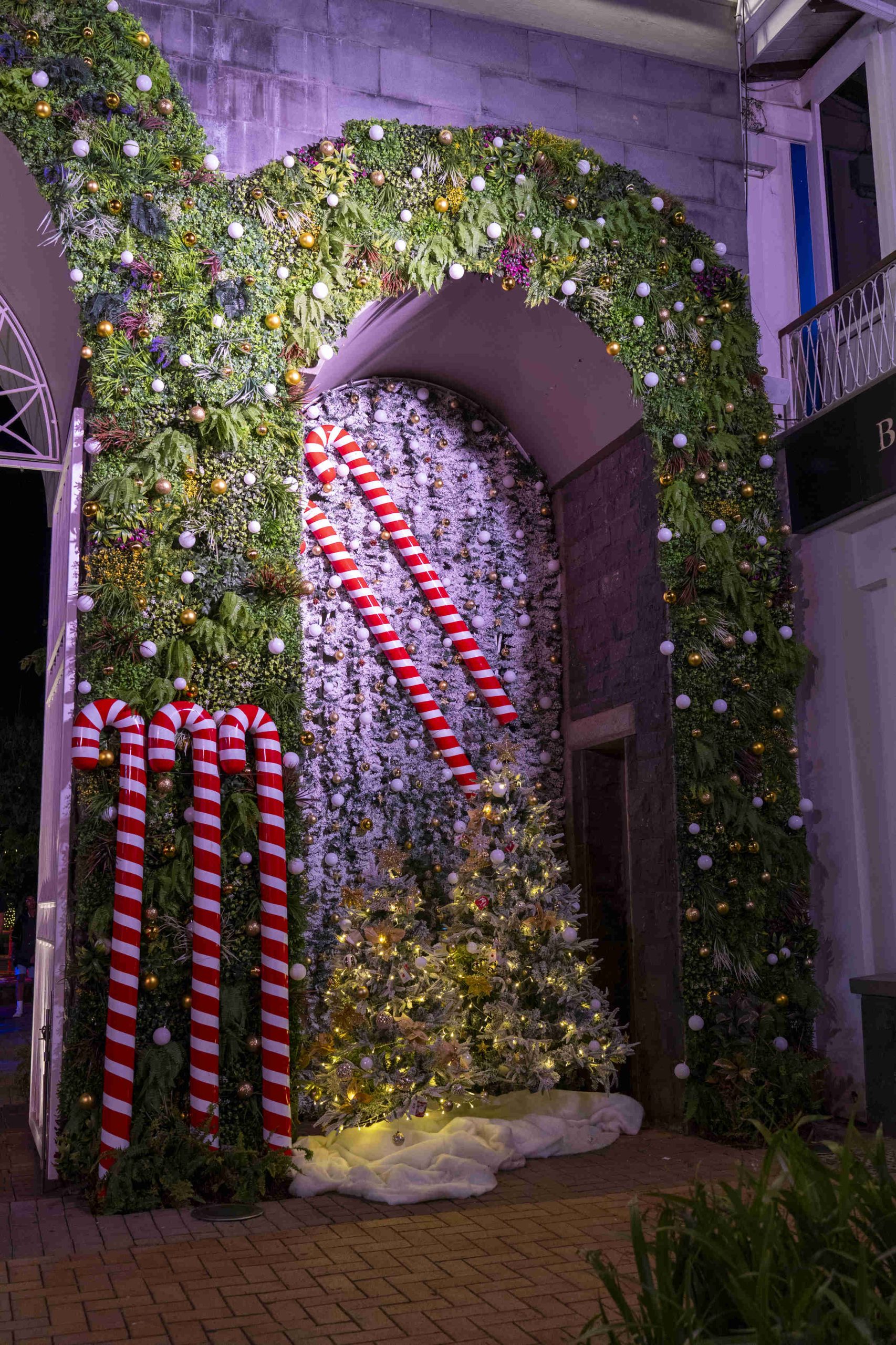 Festive archway covered in lush greenery and white ornaments, adorned with red-striped candy canes. A glowing white Christmas tree beneath adds warmth.