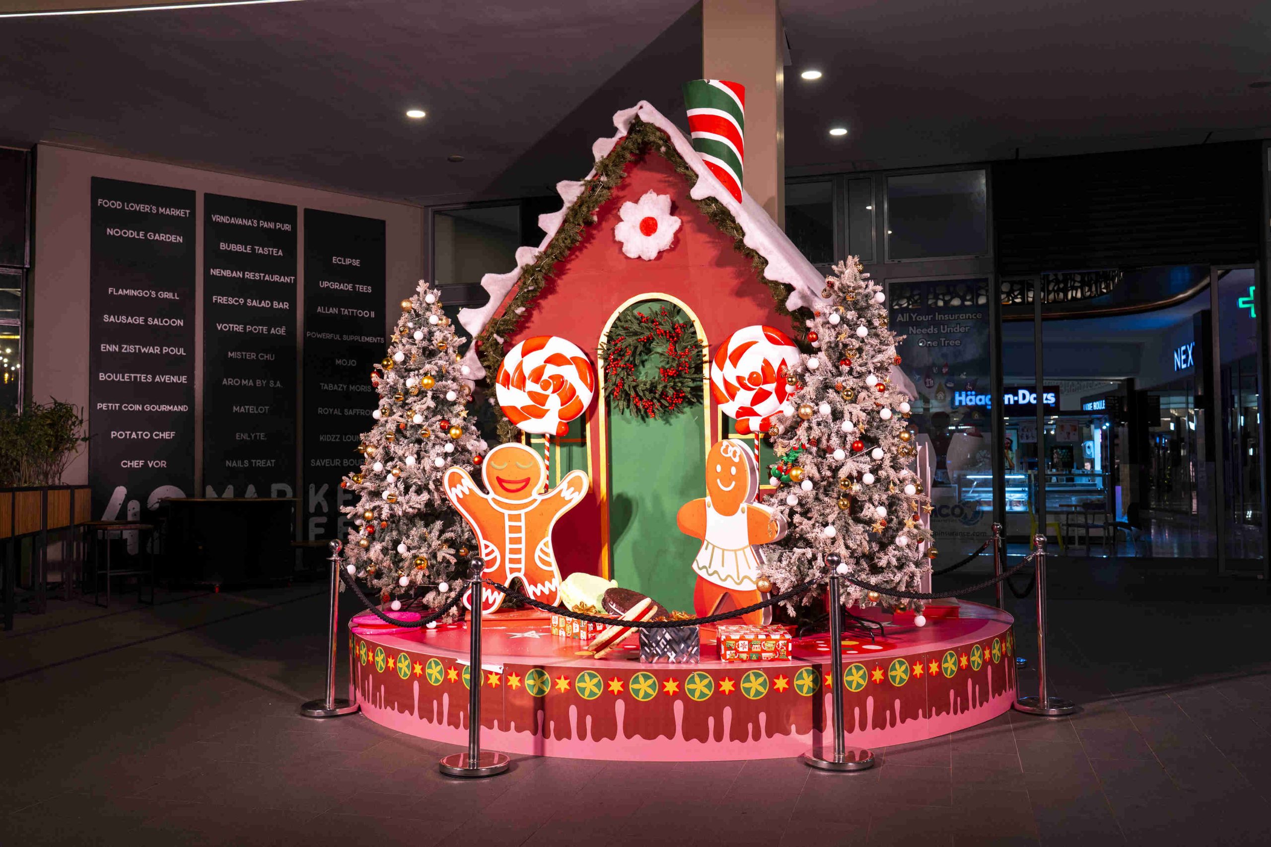 Festive gingerbread house display with candy canes, two decorated Christmas trees, and cheerful gingerbread figures. Bright, colorful holiday scene.
