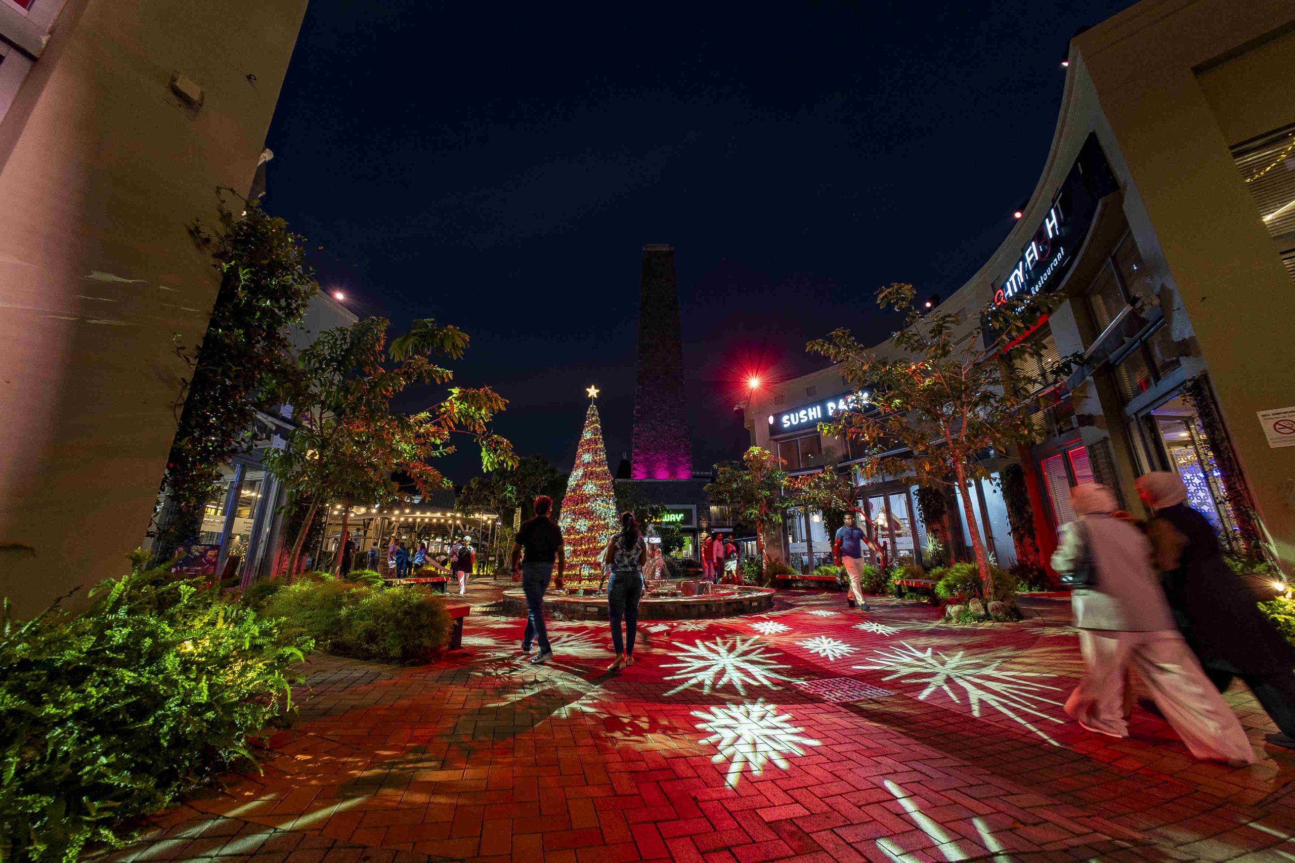 A festive scene featuring a towering Christmas tree adorned with lights and a glowing star. It's night, and artificial snow falls, creating a magical atmosphere.